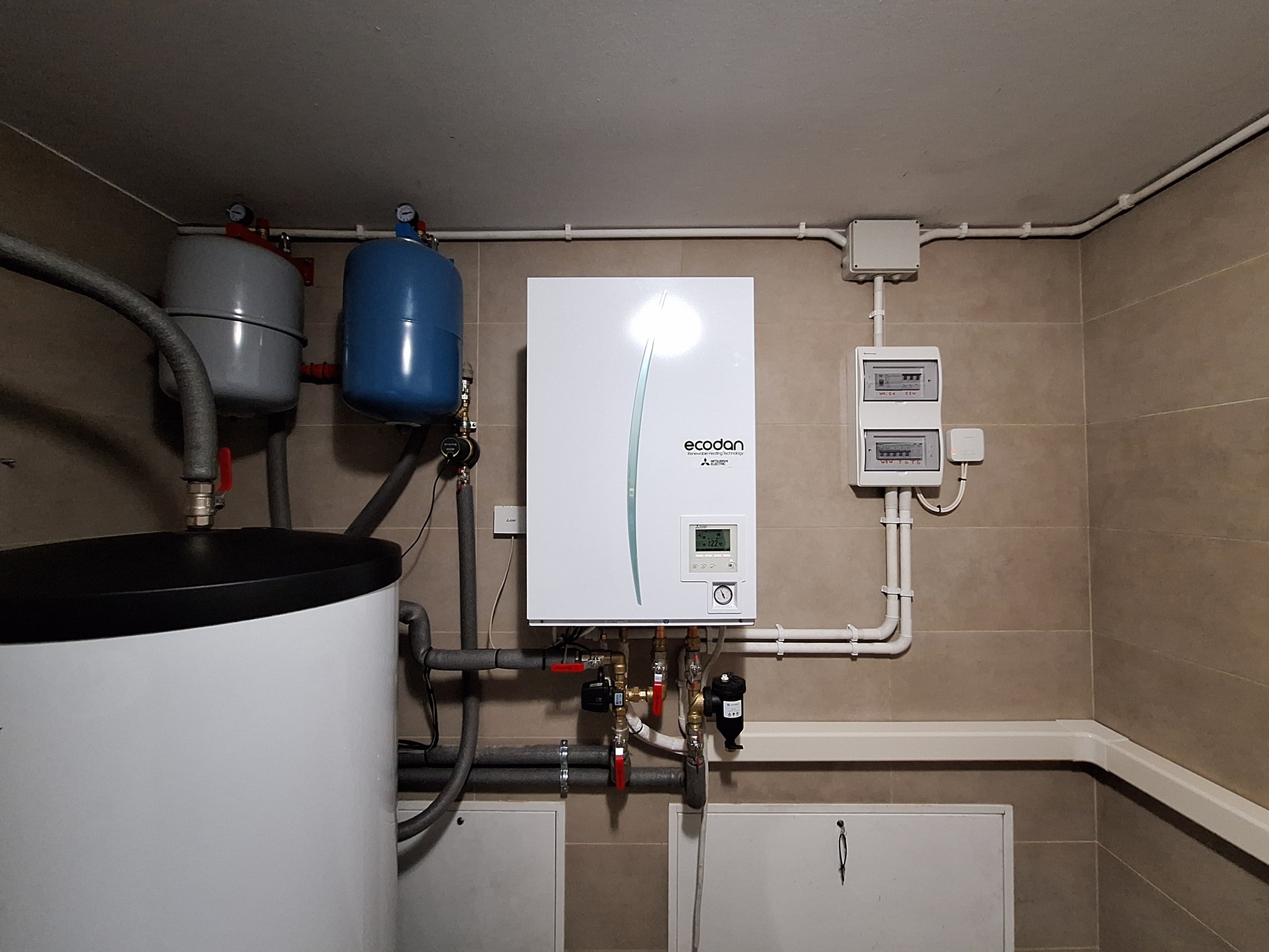 Zainstalowana biała pompa ciepła Ecodan z szarymi i niebieskimi zbiornikami wyrównawczymi w pomieszczeniu z beżowymi płytkami, widoczne rury i zabezpieczenia elektryczne.