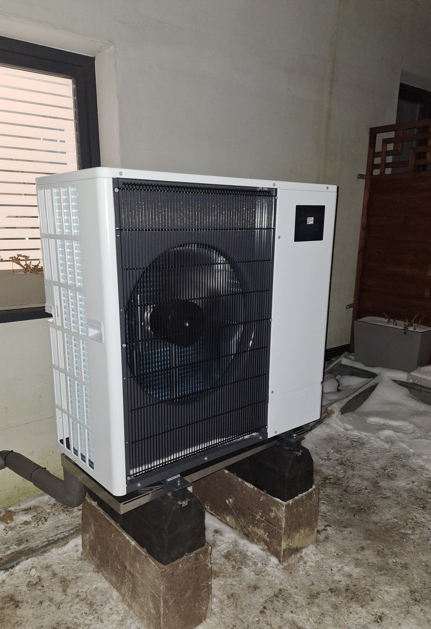 Zewnętrzna jednostka pompy ciepła powietrze-woda zamontowana na ceglanych podstawach w zimowym otoczeniu, widoczna rura odprowadzająca skropliny, na ścianie widoczne zacieki