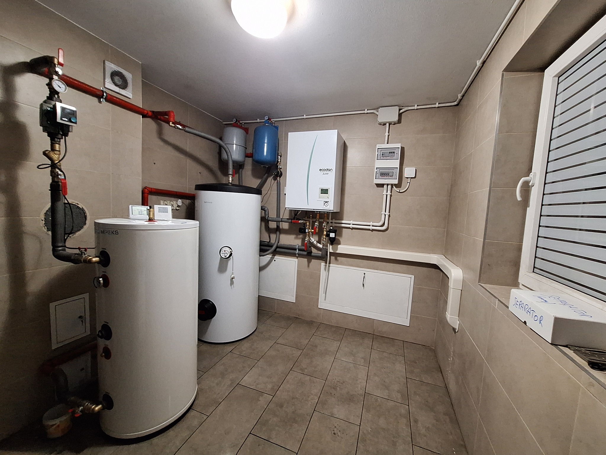 Kotłownia z dwoma białymi zbiornikami, niebieskimi naczyniami przeponowymi, kotłem gazowym, licznikiem i instalacją hydrauliczną w pomieszczeniu wyłożonym kafelkami.