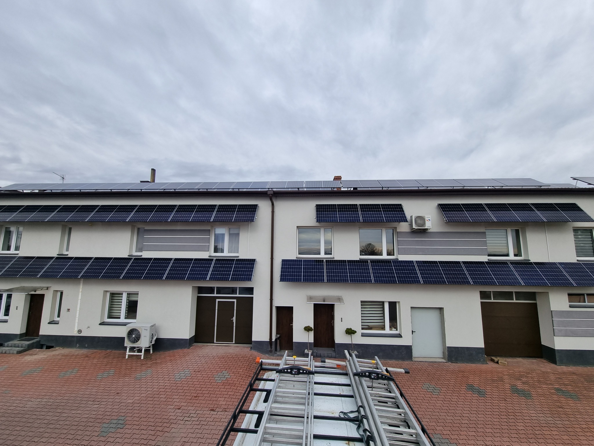 Panele fotowoltaiczne zamontowane nad oknami i na dachu segmentu domu jednorodzinnego, na pierwszym planie drabiny na przyczepce samochodowej, dodatkowo widoczny klimatyzator na zewnątrz budynku.