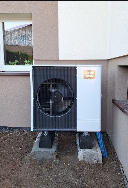 Zewnętrzna jednostka pompy ciepła zamontowana na betonowych fundamentach przy ścianie budynku z widocznym oknem i fragmentem rynny.