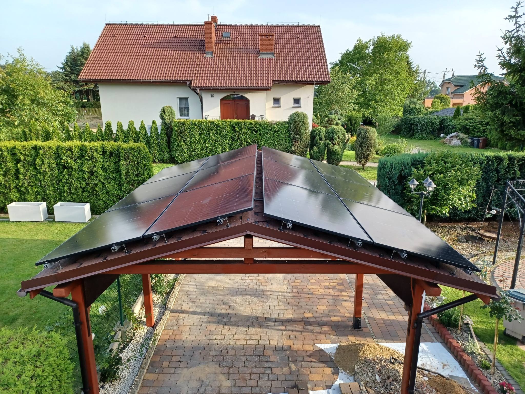 Panel słoneczny zamontowany na drewnianej wiacie garażowej, widok z góry na kostkę brukową, w tle dom jednorodzinny z czerwonym dachem.
