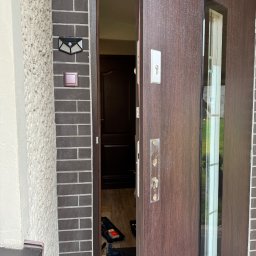 FHU EXITO Fita Arkadiusz - Częściowo otwarta, brązowa, zewnętrzna drzwi z szybą, widoczne ślady po starym zamku, narzędzia i skrzynka narzędziowa w progu.