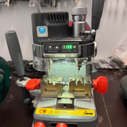 FHU EXITO Fita Arkadiusz - Maszyna do kopiowania kluczy z regulowanymi uchwytami, widocznymi w tle zawieszone klucze na ścianie.