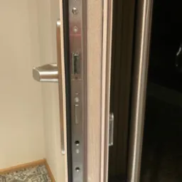 Widok na zamek i uszczelkę w otwartych drzwiach wejściowych, widoczna metalowa klamka i fragment futryny.