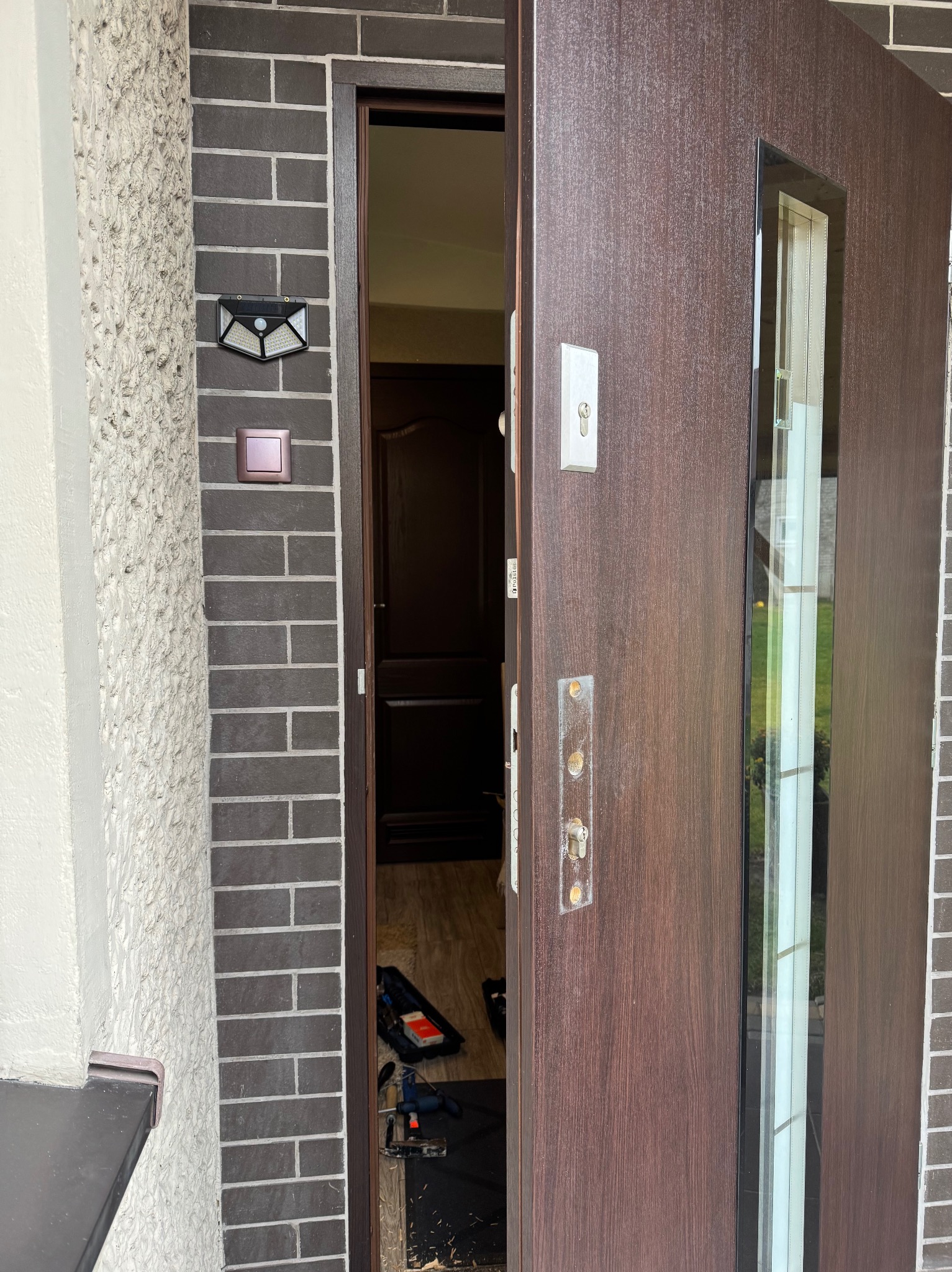 Częściowo otwarta, brązowa, zewnętrzna drzwi z szybą, widoczne ślady po starym zamku, narzędzia i skrzynka narzędziowa w progu.