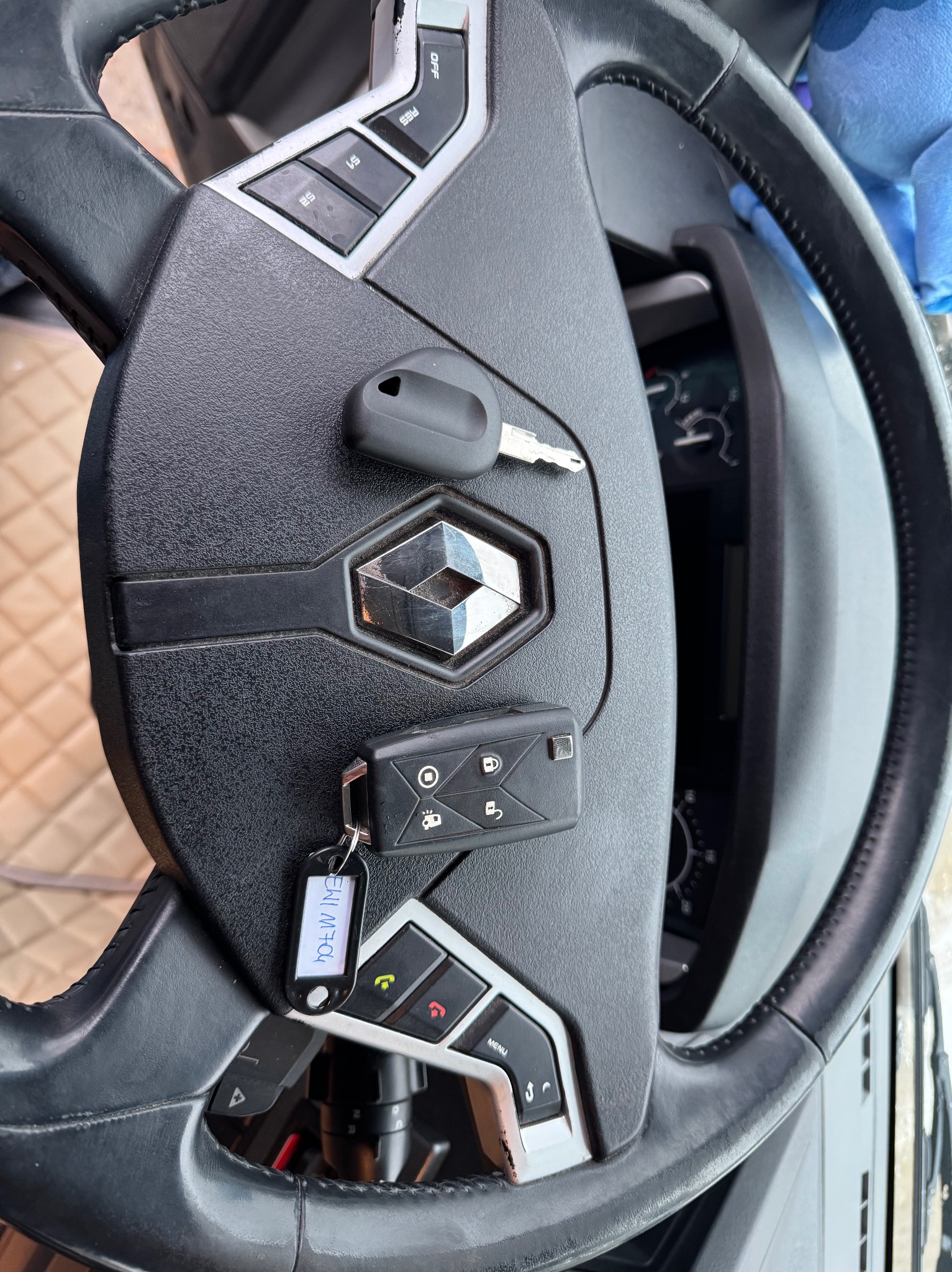 Czarna kierownica Renault z dwoma kluczykami, jeden z pilotem i brelokiem z napisem EWI M7Q4, leżącymi na centralnej części z logo.