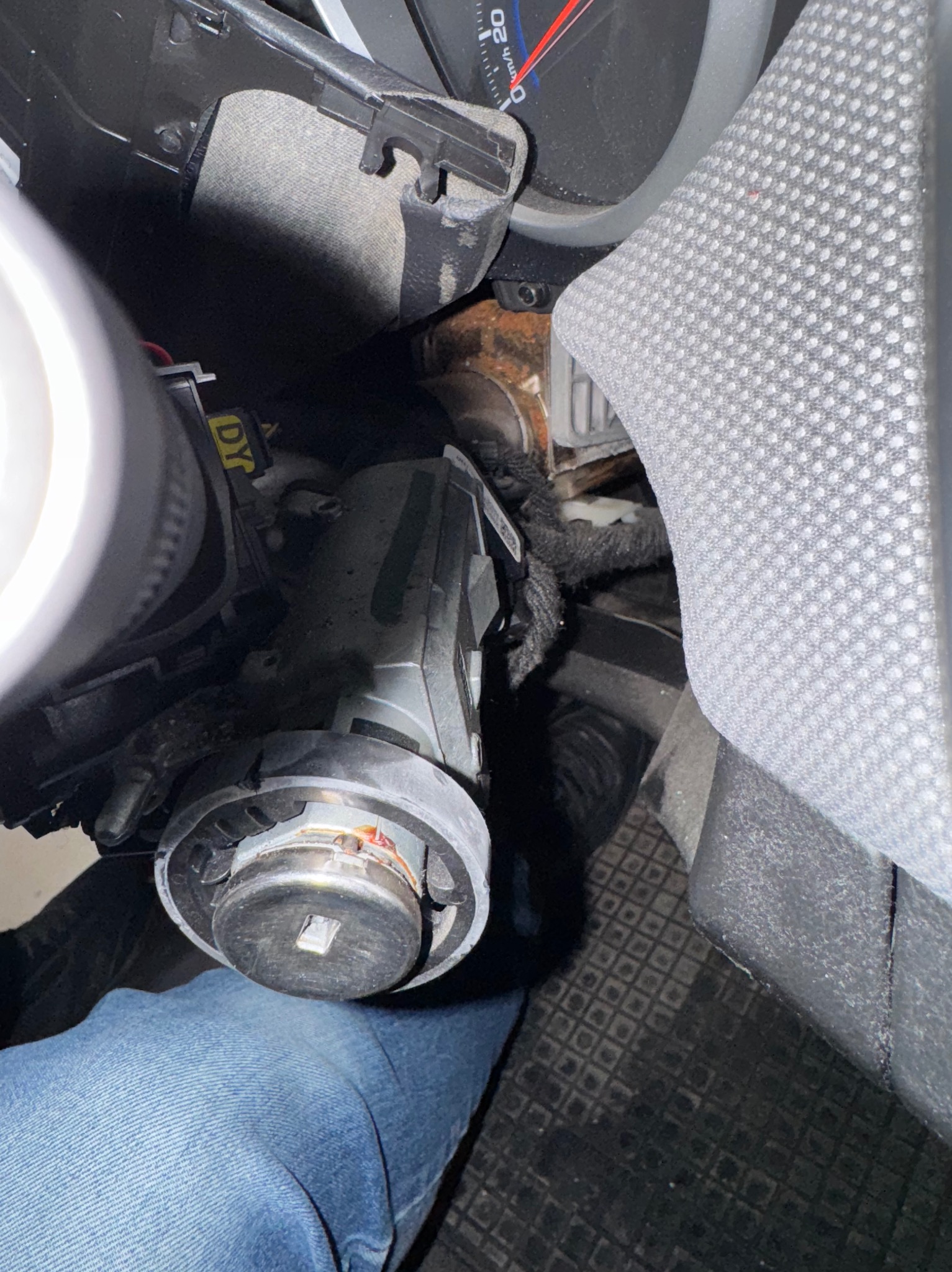 Widok na stacyjkę samochodową podczas naprawy, widoczne przewody i fragment deski rozdzielczej, jeansy osoby wykonującej usługę.