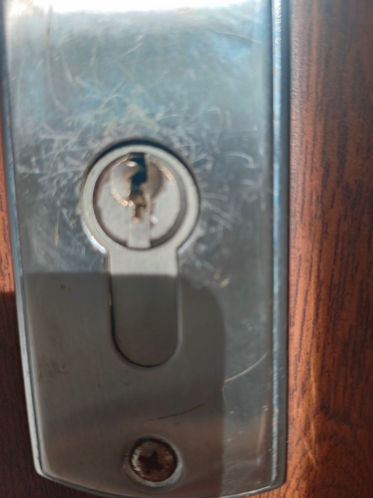 Szczegółowe zbliżenie wkładki zamka w drzwiach z widocznymi śladami użytkowania i zarysowaniami na metalowej obudowie, osadzonej w drewnianej ramie.