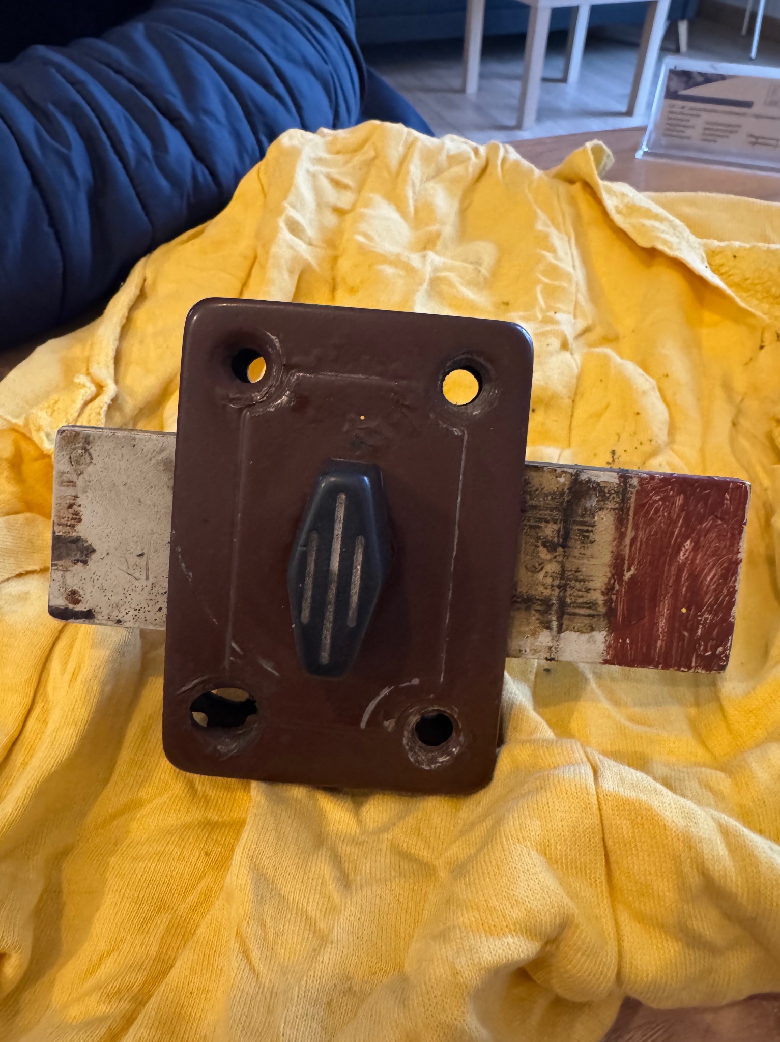 Stary, brązowy zamek do drzwi z czarną klamką, leżący na żółtej szmatce, prawdopodobnie przed montażem lub po demontażu.