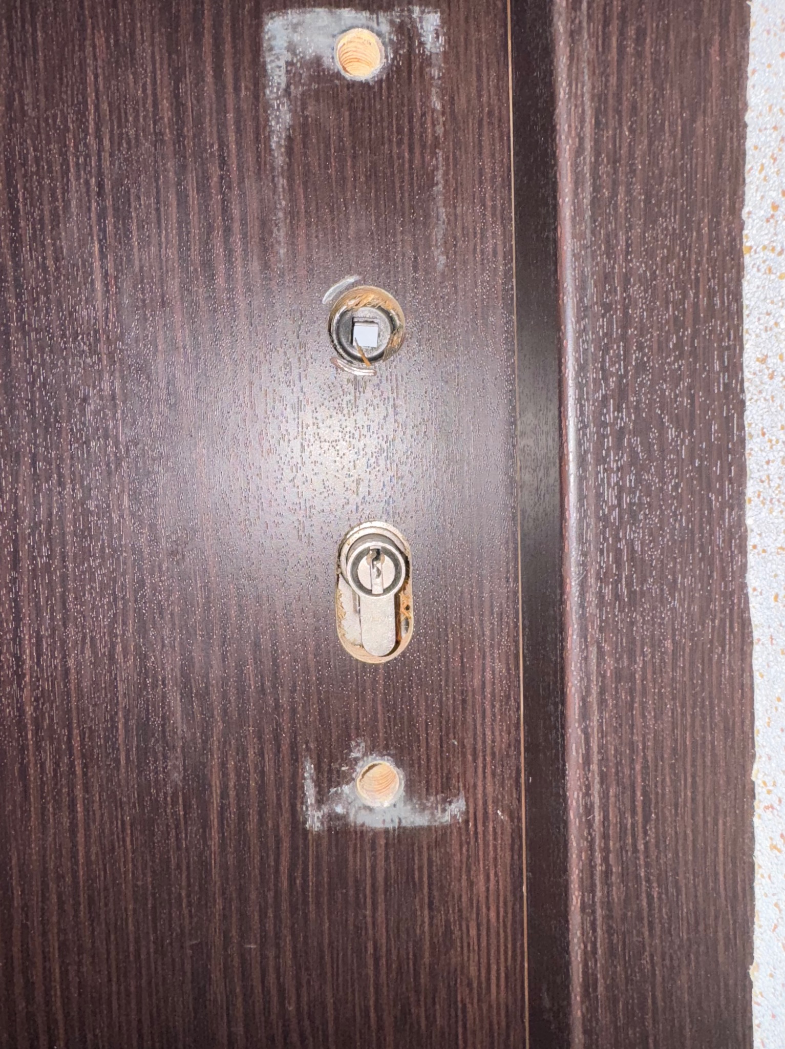 Brązowe drzwi wejściowe z widocznymi otworami po zamku i śladami po montażu, z zamontowanym zamkiem tymczasowym.
