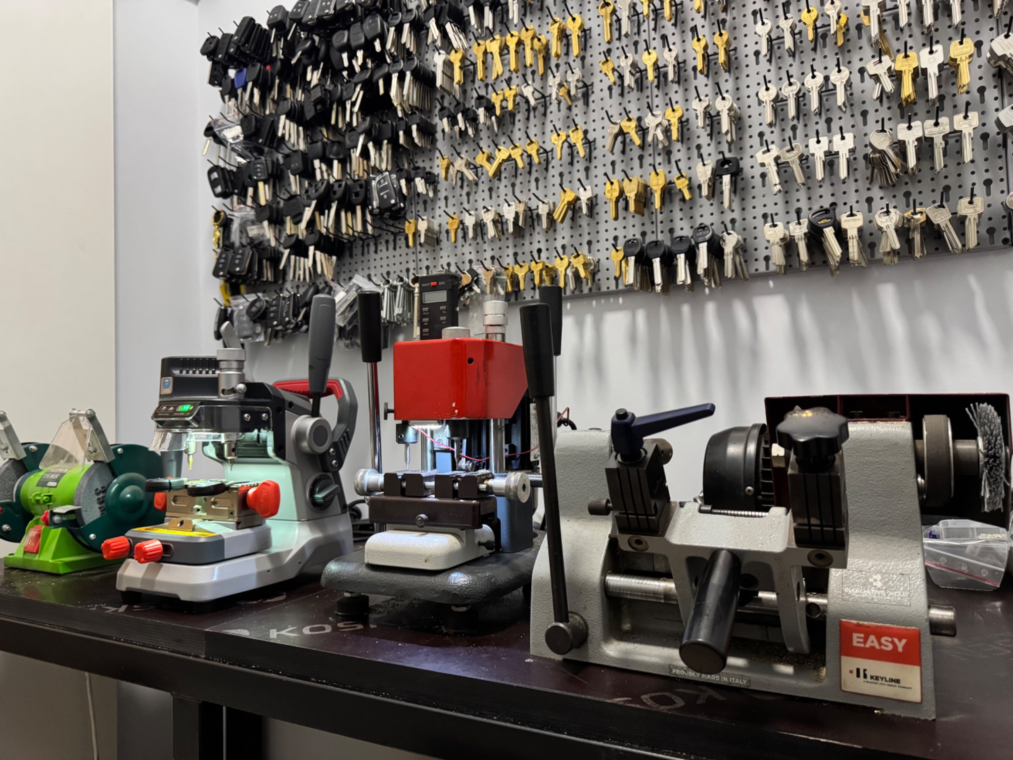 Warsztat ślusarski: Rząd maszyn do kopiowania kluczy na stole z widoczną ścianą pełną różnorodnych kluczy na perforowanej płycie.
