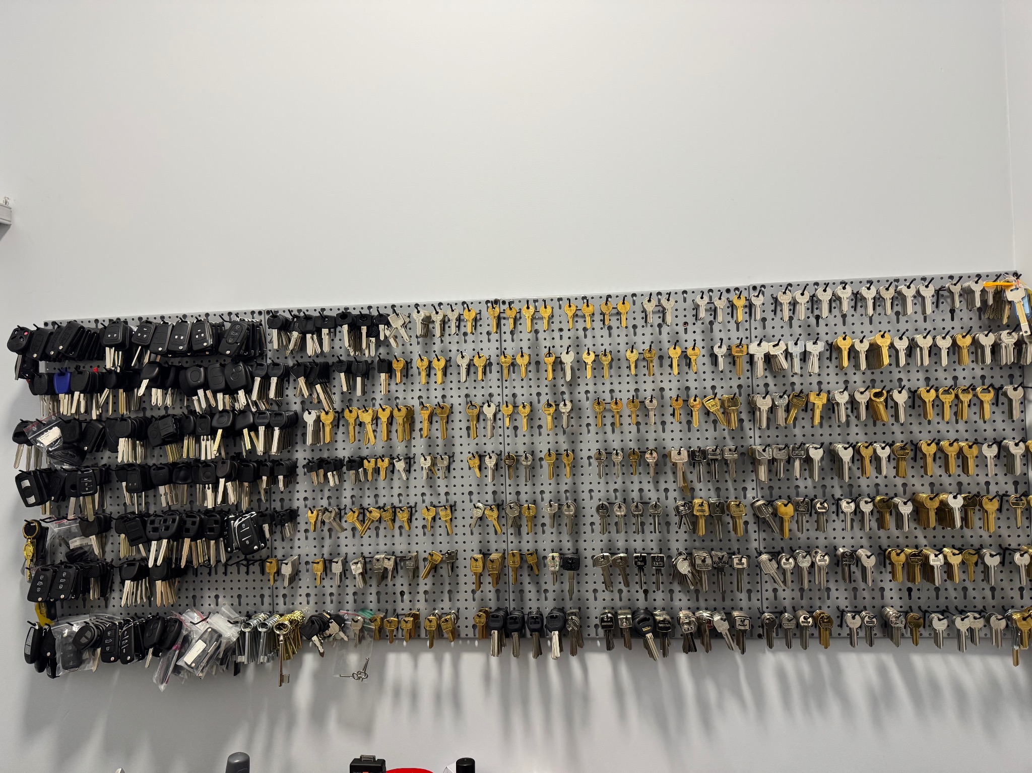 Ściana warsztatu ślusarskiego z ekspozycją różnorodnych kluczy, w tym kluczy samochodowych z pilotami, zawieszonych na perforowanej płycie.