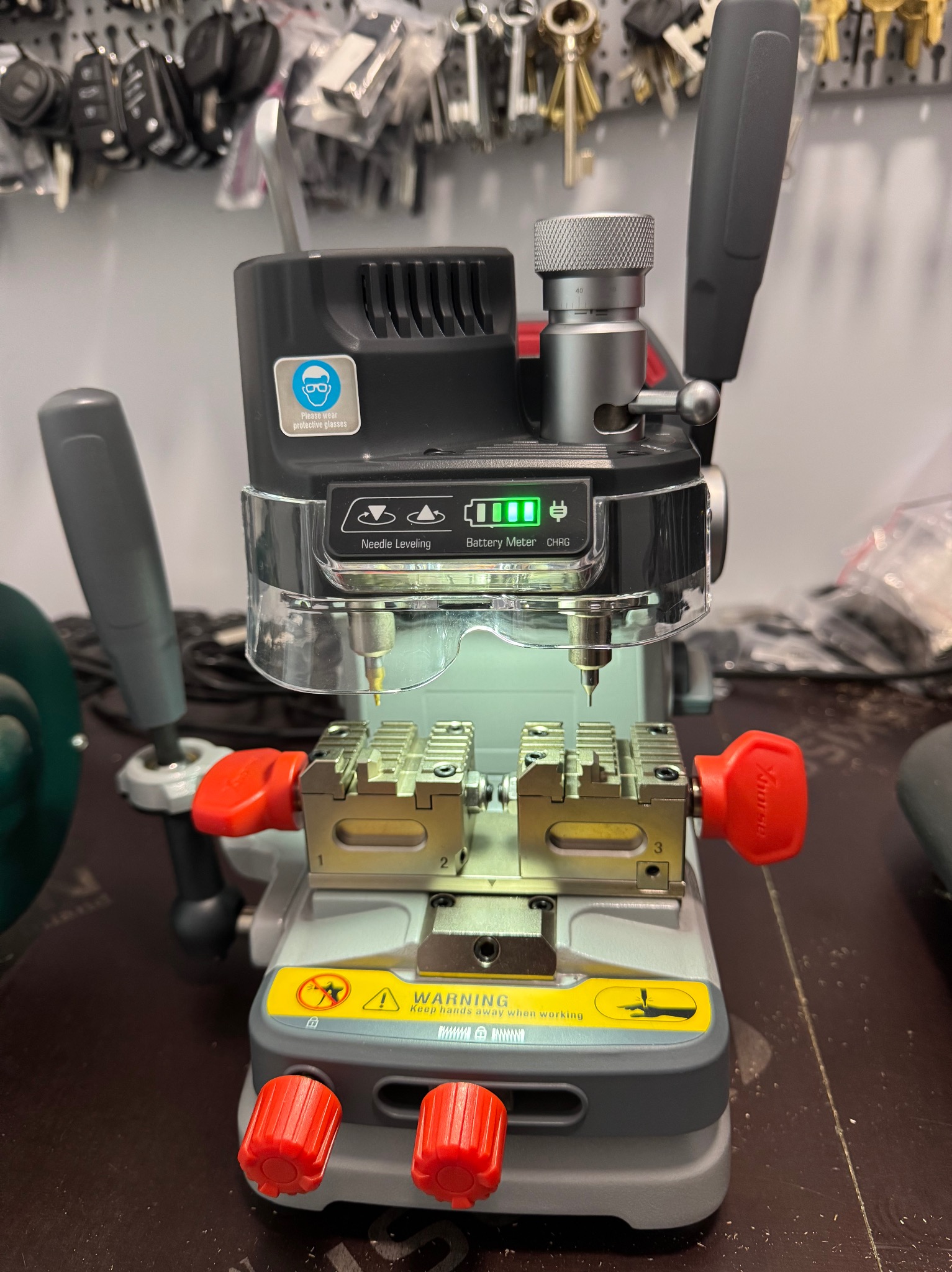 Maszyna do kopiowania kluczy z regulowanymi uchwytami, widocznymi w tle zawieszone klucze na ścianie.