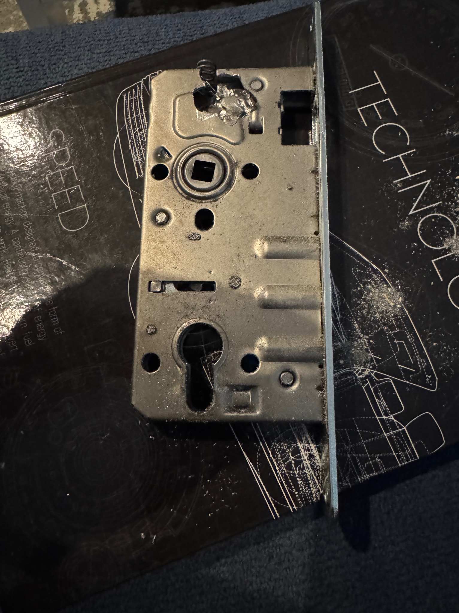 Metalowa blacha zamka drzwiowego, widoczne ślady obróbki, leżąca na czarnej książce z napisem TECHNOLOGY i SPEED.