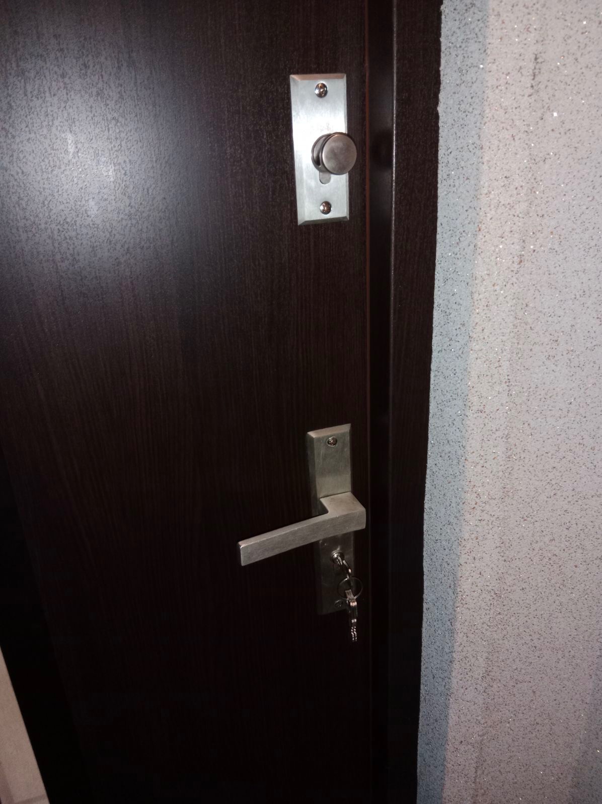 Ciemne, drewnopodobne drzwi wejściowe z zamontowanym srebrnym zamkiem z klamką i kluczami w zamku oraz dodatkowym zamkiem gałkowym, widoczne na tle jasnej ściany o chropowatej strukturze.