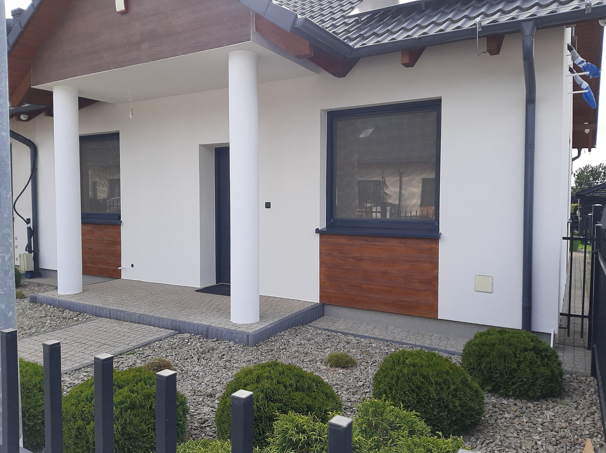 Elewacja domu z białymi ścianami, drewnianymi panelami pod oknami, kolumnami przy wejściu i szarą kostką brukową na podjeździe, otoczona zielonymi krzewami i żwirem.