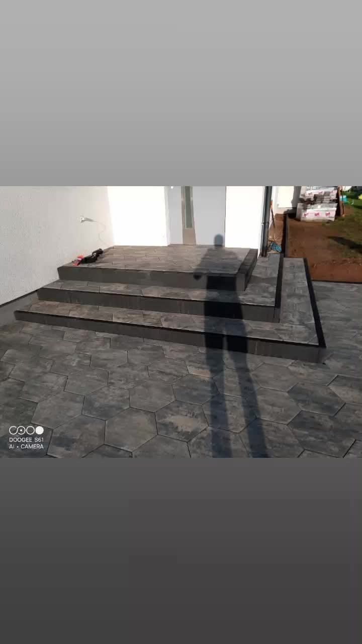 Schody wejściowe do domu wyłożone szarą, heksagonalną kostką brukową, z widocznym cieniem osoby fotografującej i paletą kostki w tle.