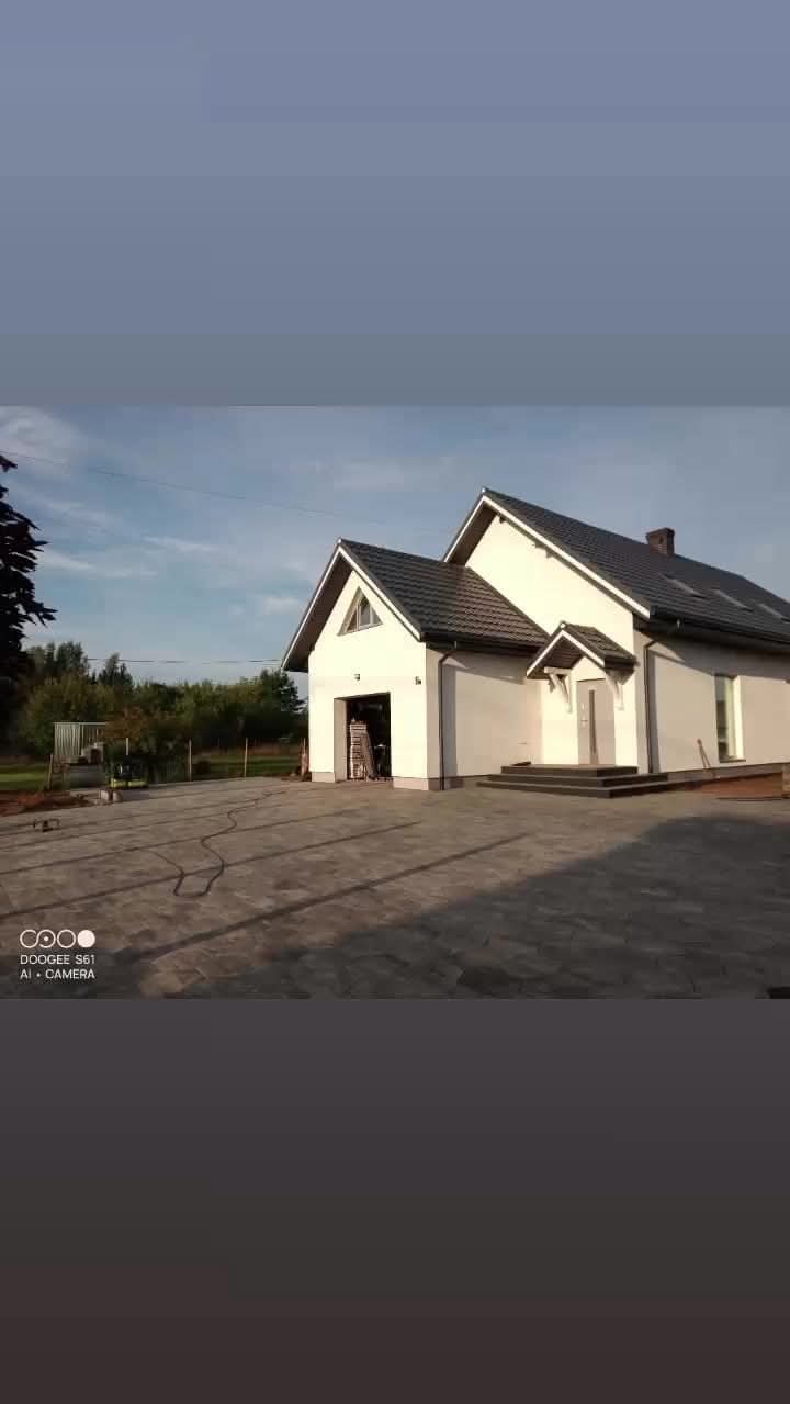 Nowo ułożona, szara kostka brukowa przed nowoczesnym domem z białymi ścianami i ciemnym dachem, widoczne ślady po kablach i lekki bałagan.
