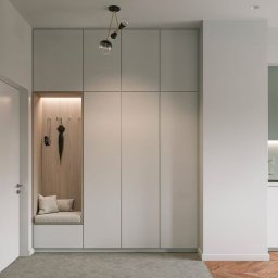 MEBLE od SERCA - Nowoczesna szafa wnękowa z siedziskiem i wieszakami na ubrania, oświetlona lampą w stylu retro, na tle szarej podłogi i drewnianego parkietu.