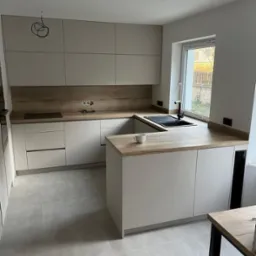 Jasna kuchnia z drewnianym blatem, wyspą i czarnym zlewem, widok na okno z zewnątrz. W tle niedokończone podłączenie oświetlenia.