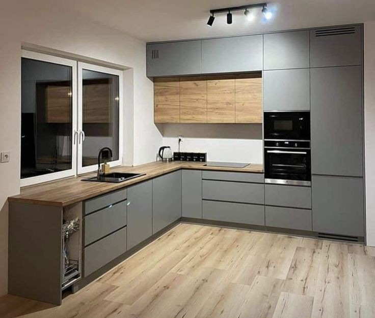 Szara, narożna zabudowa kuchenna z drewnianym blatem i górnymi szafkami o fakturze drewna, z oknem po lewej stronie i drewnianą podłogą.