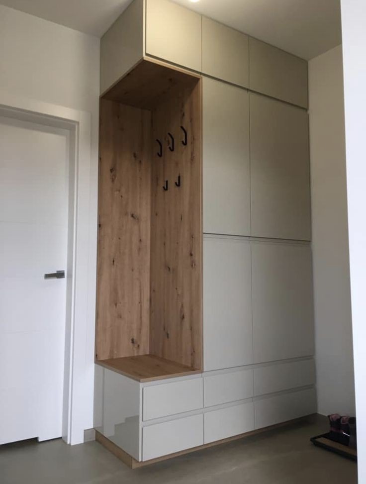 Wnęka z wieszakami i półką, wkomponowana w zabudowę meblową z szufladami i szafkami w jasnych odcieniach, obok białe drzwi.