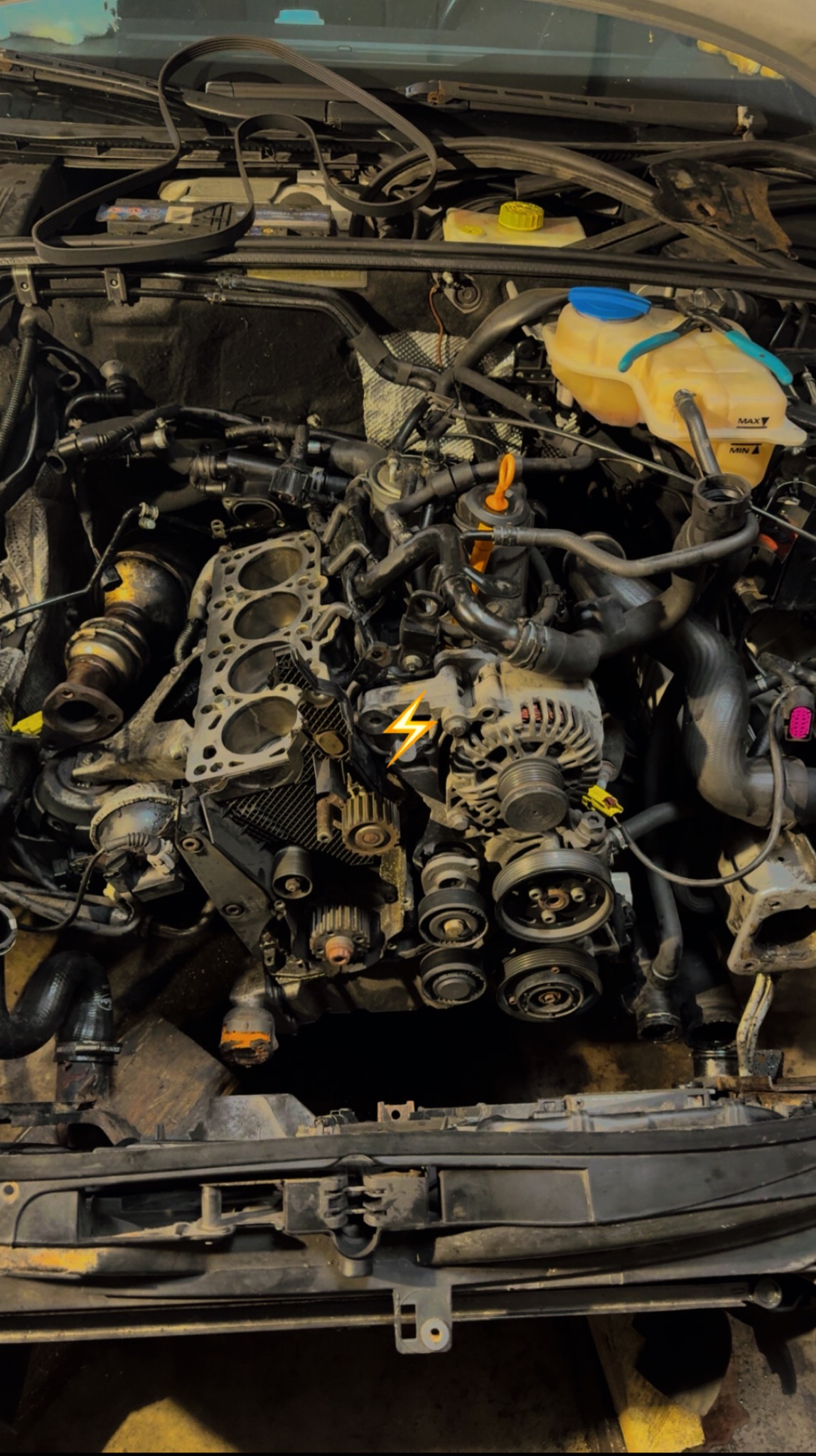 Silnik samochodowy w trakcie naprawy, widoczny zdemontowany blok cylindrów, elementy układu rozrządu i osprzęt, w tle zbiorniczki płynów eksploatacyjnych.