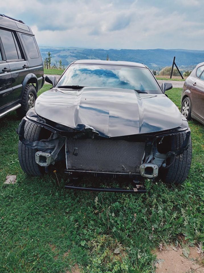 Uszkodzony czarny samochód sportowy z przodu, brak elementów karoserii, widoczny radiator, na trawie, w tle górzysty krajobraz.