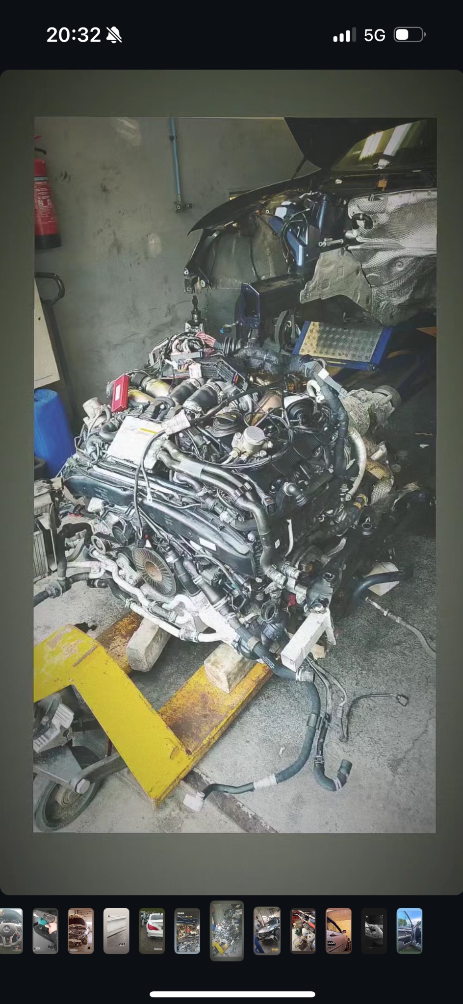 Wyjęty silnik samochodowy umieszczony na żółtym wózku warsztatowym, widoczny fragment karoserii samochodu na podnośniku warsztatowym w tle, ściana z widocznym sprzętem gaśniczym.