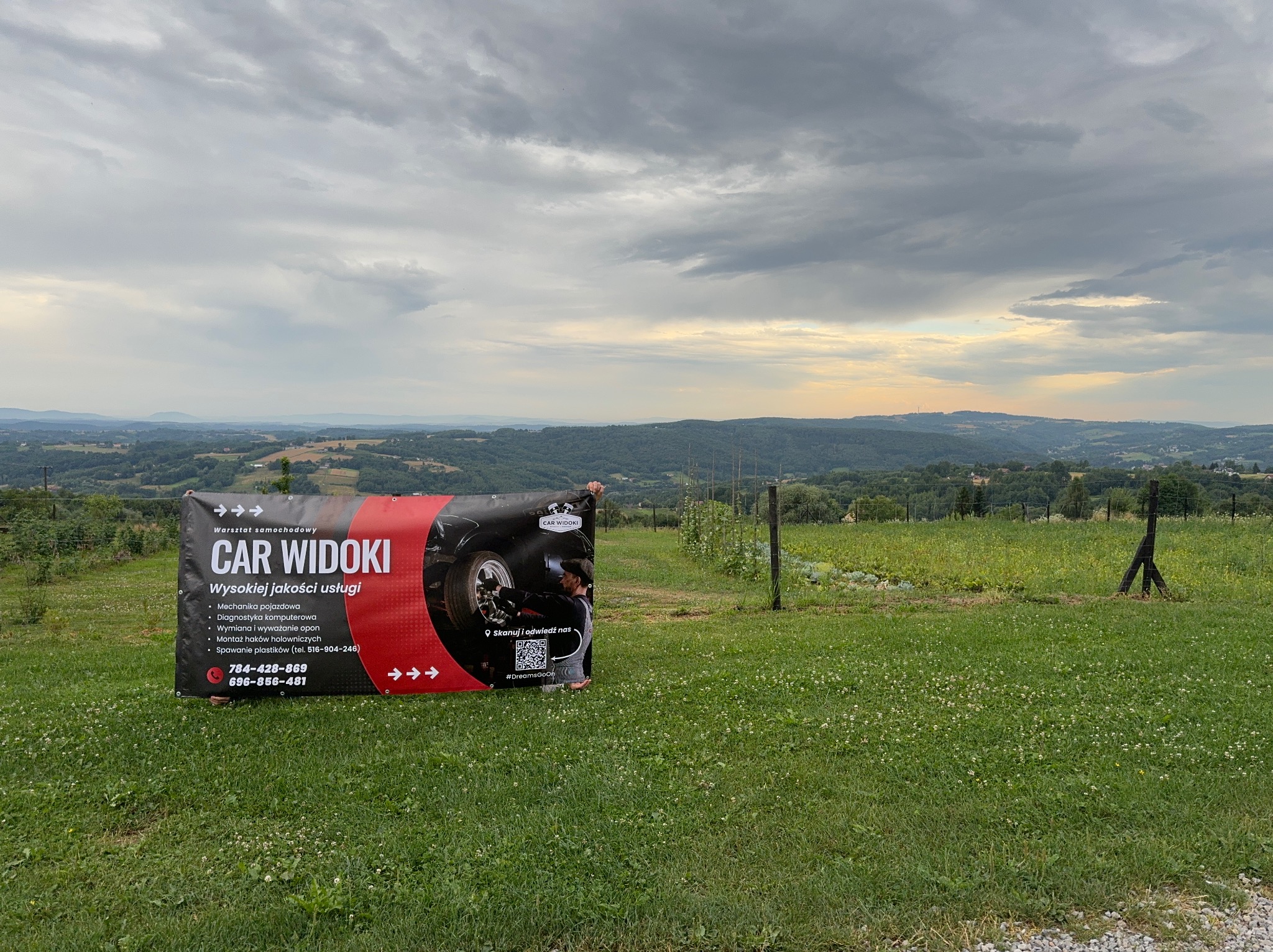 Baner reklamowy warsztatu samochodowego 'Car Widoki' na tle zielonego pola i malowniczego krajobrazu z widokiem na góry pod pochmurnym niebem.