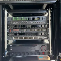 Szafa rack z zainstalowanymi urządzeniami sieciowymi: patch panel, switche, rejestrator BCS z listwą zasilającą, widoczne kable i elementy montażowe.
