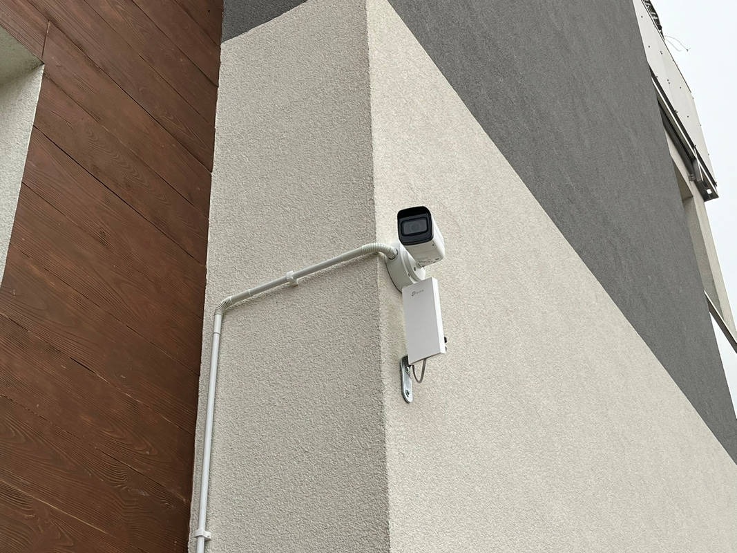 Kamera monitoringu TP-Link zamontowana na elewacji budynku, z widocznymi przewodami w białej rurce ochronnej, na tle beżowej i szarej ściany z elementami drewnianymi.