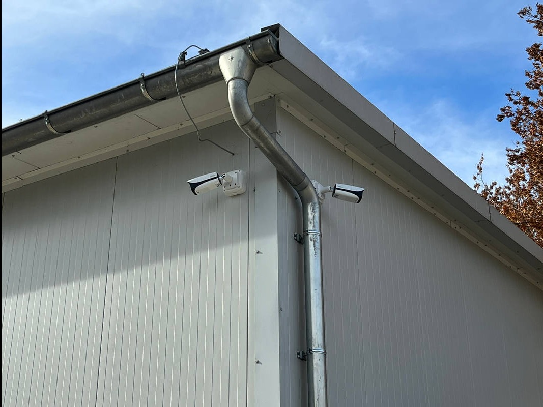 Dwie kamery monitoringu zamontowane na zewnętrznej ścianie budynku z blachy trapezowej, obok rynny spustowej, widok pod kątem, błękitne niebo w tle.