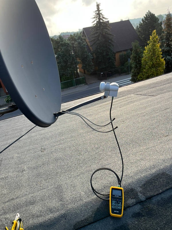 Antena satelitarna na dachu podczas pomiarów sygnału miernikiem, w tle domy i drzewa.