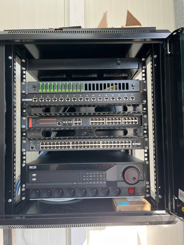 Szafa rack z zainstalowanymi urządzeniami sieciowymi: patch panel, switche, rejestrator BCS z listwą zasilającą, widoczne kable i elementy montażowe.