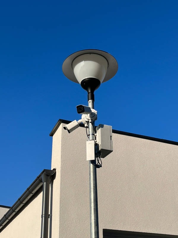 Słup oświetleniowy z zamontowanymi kamerami monitoringu zewnętrznego i puszką instalacyjną na tle budynku z rynną.