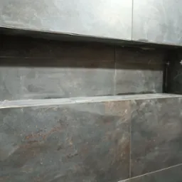 Ściana z ciemnych, matowych płytek imitujących metal z wnęką wykończoną metalową listwą, w trakcie montażu widoczne krzyżyki dystansowe.