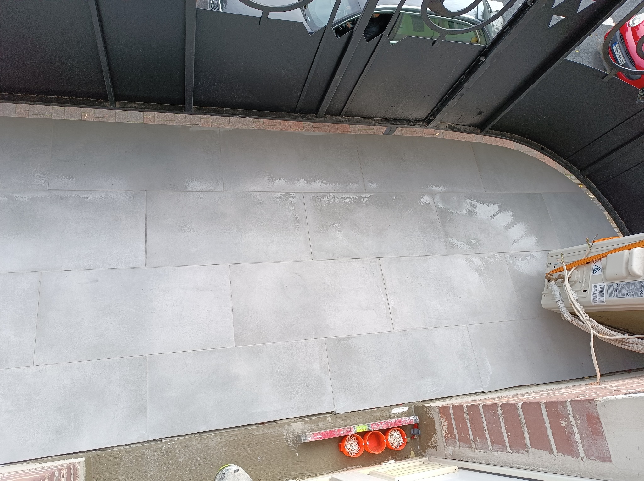 Świeżo ułożone, duże, szare płytki na ścianie zewnętrznej pod zadaszeniem, widoczne przygotowane puszki elektryczne i poziomica.