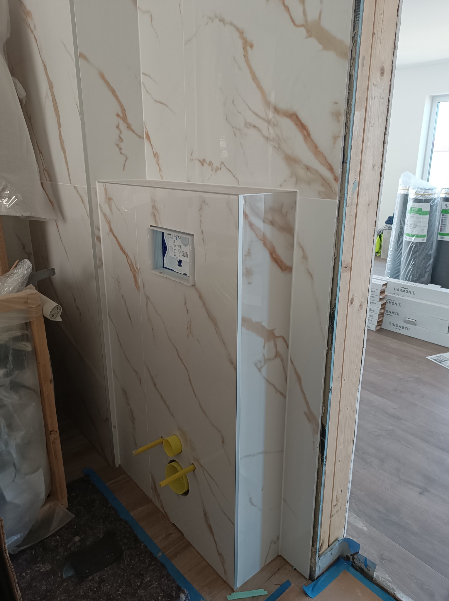 Ściana łazienki w trakcie wykańczania, widoczne płytki imitujące marmur, stelaż podtynkowy WC z żółtymi rurami doprowadzającymi wodę, zabezpieczone podłoże folią malarską, w tle opakowania płytek.