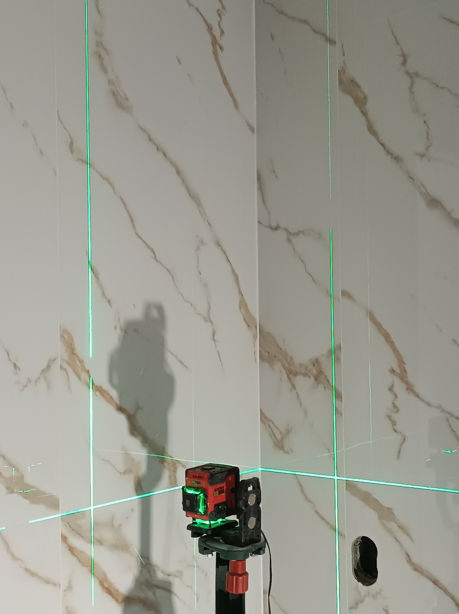 Laser krzyżowy wyznaczający pion i poziom na płytkach imitujących marmur, widoczny otwór na instalację.