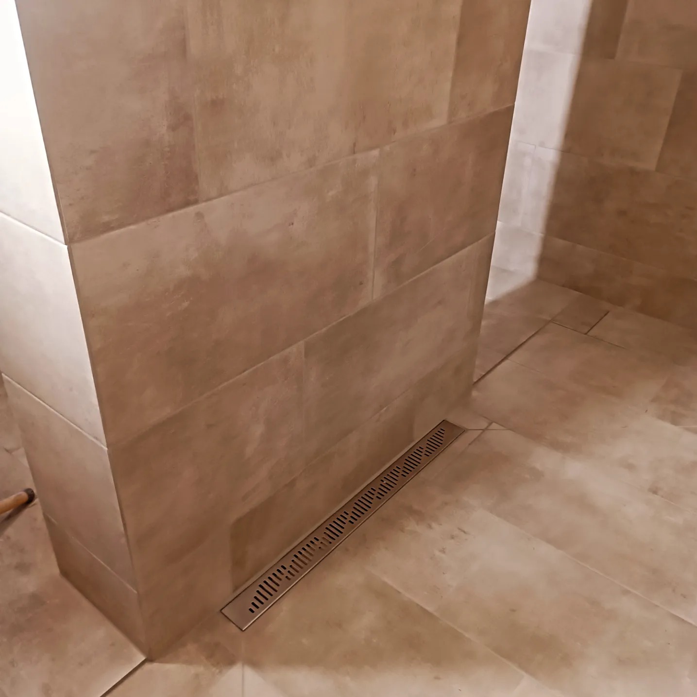 Beżowe płytki ścienne i podłogowe w łazience z odpływem liniowym w rogu kabiny prysznicowej.