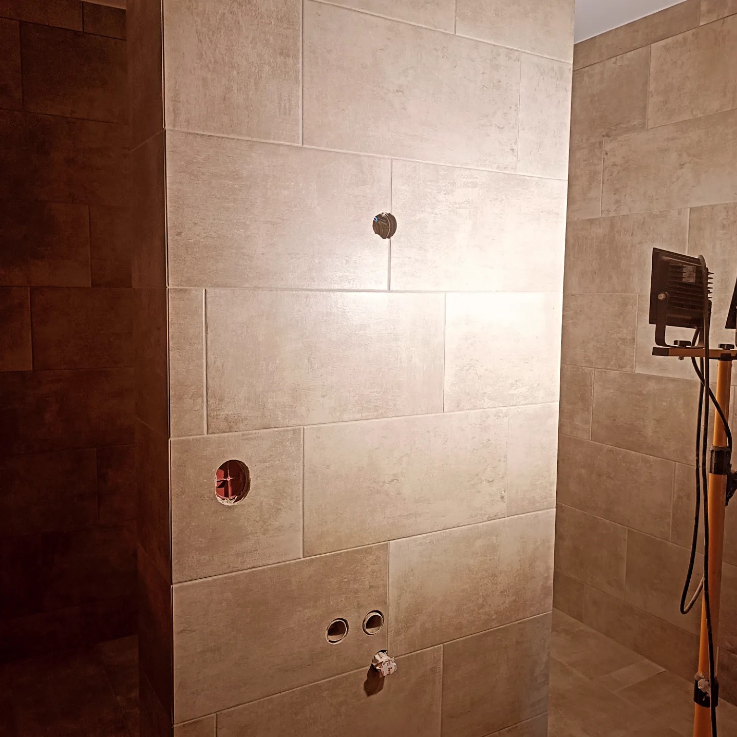 Ściana łazienki wyłożona beżowymi płytkami w trakcie montażu, widoczne otwory pod instalacje, oświetlenie budowlane po prawej stronie.