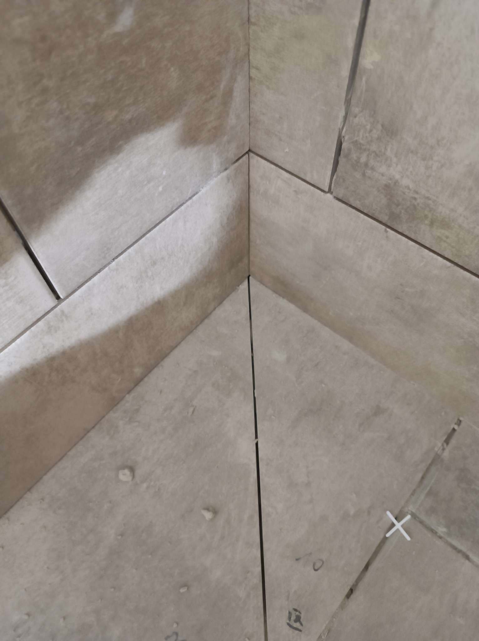 Ukośne ułożenie beżowych płytek w rogu pomieszczenia, widoczne krzyżyki dystansowe i fuga w trakcie schnięcia.