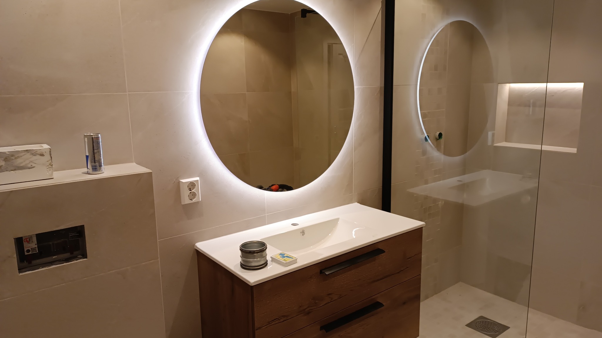 Nowoczesna łazienka z beżowymi płytkami, okrągłym lustrem z podświetleniem LED, drewnianą szafką pod umywalką i kabiną prysznicową ze szklaną ścianką.