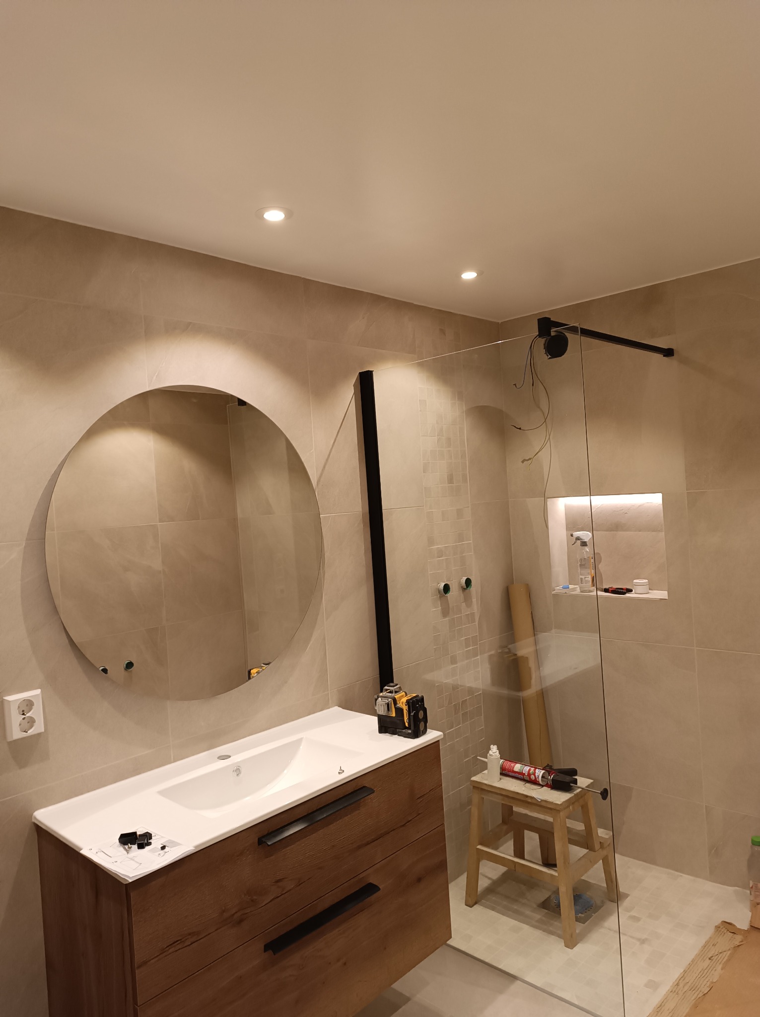 Nowoczesna łazienka w trakcie wykańczania: okrągłe lustro nad drewnianą szafką z umywalką, kabina prysznicowa ze szklaną ścianą i czarnymi elementami, widoczne narzędzia i materiały budowlane...