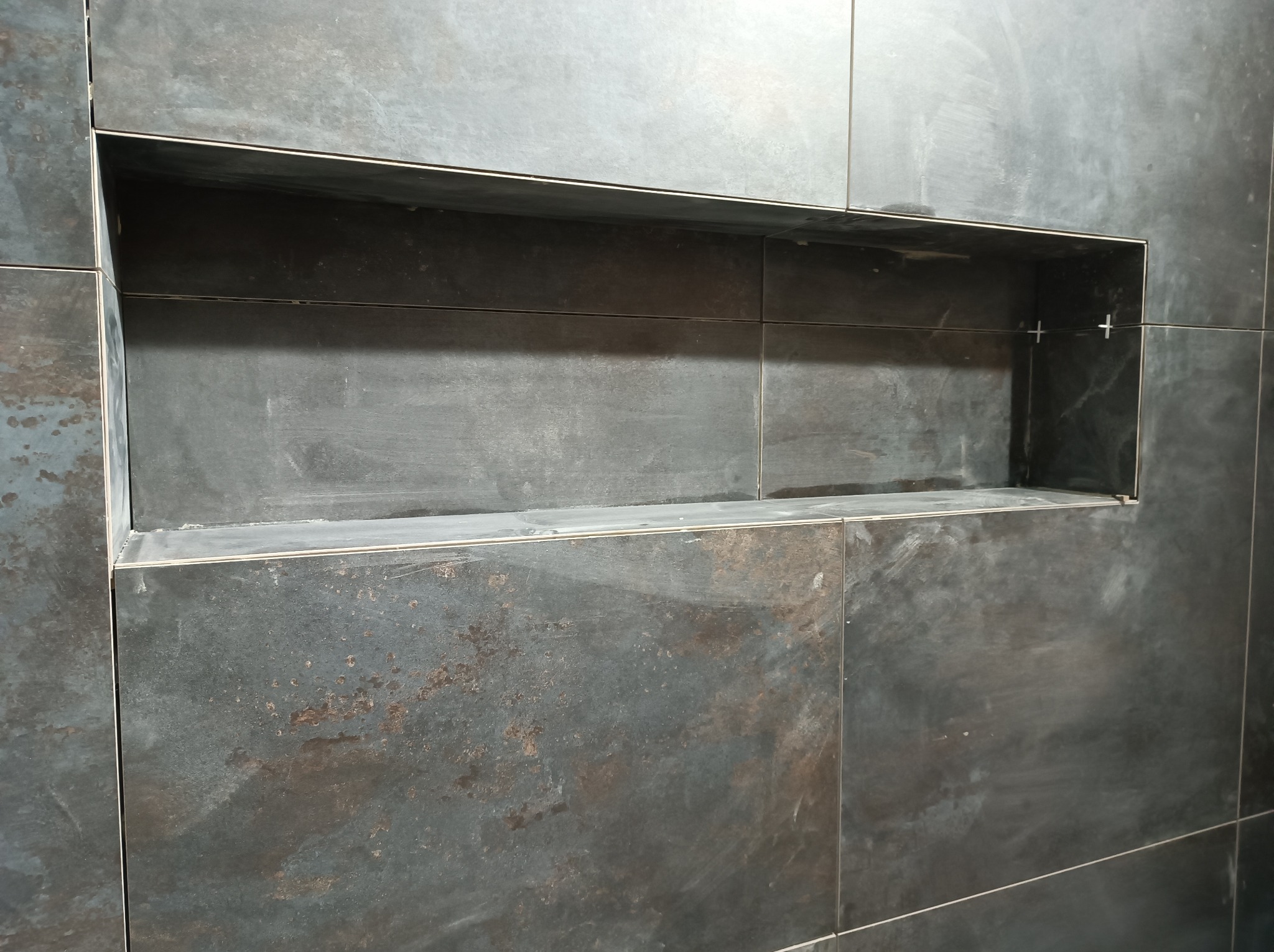 Ściana z ciemnych, matowych płytek imitujących metal z wnęką wykończoną metalową listwą, w trakcie montażu widoczne krzyżyki dystansowe.