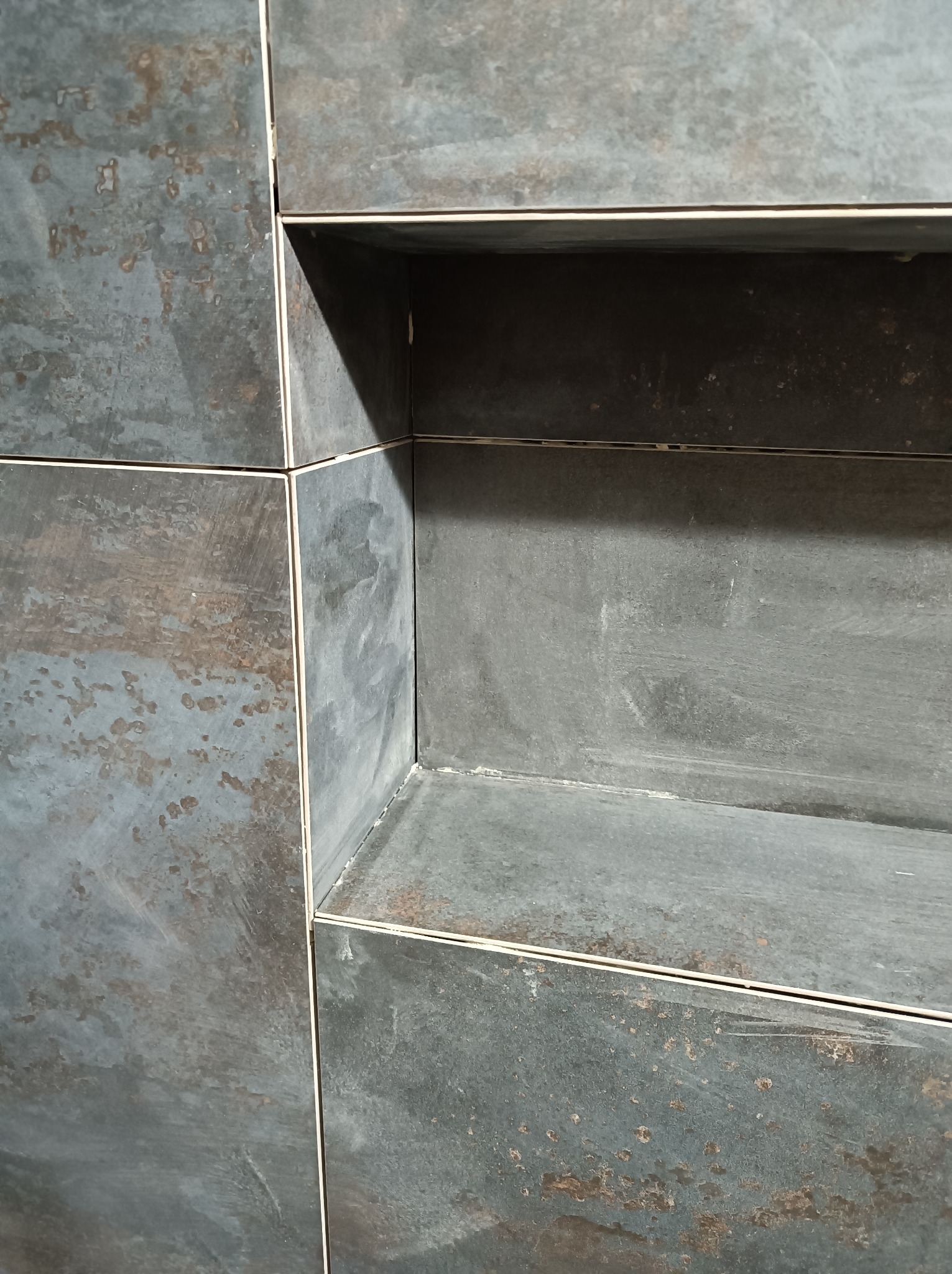 Narożne ujęcie wnęki wyłożonej ciemnymi, metalizowanymi płytkami w łazience, z widocznymi fugami w kolorze ecru.