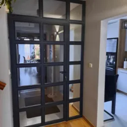 Szklana ściana działowa w metalowej ramie, z widocznym fragmentem schodów i wnętrzem domu, oświetlonym lampą sufitową.