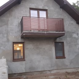 KB SAFETY Kamil Kałużny - Szary dom z brązowym balkonem i oknami, widoczny mężczyzna przy ogrodzeniu, w tle zieleń.